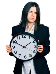 Poster - Young brunette woman with blue eyes holding big clock skeptic and nervous, frowning upset because of problem. negative person.