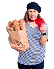 Wall Mural - Young beautiful blonde woman wearing french beret holding bag with bread annoyed and frustrated shouting with anger, yelling crazy with anger and hand raised