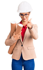 Poster - Young beautiful woman wearing architect hardhat and glasses holding blueprints pointing with finger to the camera and to you, confident gesture looking serious