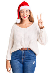 Canvas Print - Beautiful young woman wearing christmas hat smiling with happy face winking at the camera doing victory sign. number two.