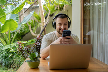 Young happy creative freelancer man in casual clothes working in coworking space wearing headphones on laptop. Remote work on the tropical island of Bali, relocation