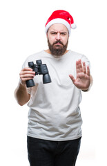 Sticker - Young caucasian hipster man wearing christmas hat looking though binoculars over isolated background with open hand doing stop sign with serious and confident expression, defense gesture