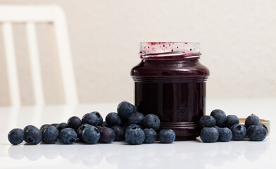 Glass jar of tasty sweet blueberry jam and fresh ripe berries on white surface..