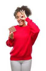 Poster - Young afro american woman eating chocolate bar over isolated background with happy face smiling doing ok sign with hand on eye looking through fingers