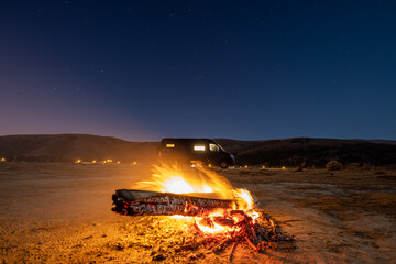 Poster - Large campfire in the foreground in the early evening with a campervan in the background.