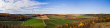 Aerial Drone Panoramic View Of Agricultural Fields Bordered By Woods With Autumn Colours And Wind Turbines In The Background Under A Semi-clear Sky In Lower Austria.