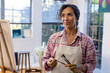 © Wavebreak Media - Happy biracial woman wearing apron holding brushes and looking away in home studio