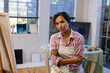 © Wavebreak Media - Portrait of happy biracial woman wearing apron holding brushes in home studio