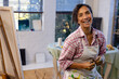 © Wavebreak Media - Portrait of happy biracial woman wearing apron holding brushes in home studio