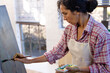© Wavebreak Media - Biracial woman wearing apron painting on easel using brush in home studio