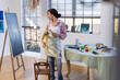 © Wavebreak Media - Biracial woman wearing apron painting on easel using brush in home studio