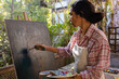 © Wavebreak Media - Biracial woman wearing apron painting on easel using brush and colour palette in garden