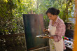 © Wavebreak Media - Biracial woman wearing apron painting on easel using brush and colour palette in garden