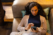 © Wavebreak Media - Biracial woman wearing headphones lying on sofa and using smartphone in living room
