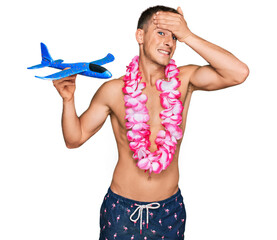 Poster - Young blond man wearing swimsuit and hawaiian lei holding airplane toy stressed and frustrated with hand on head, surprised and angry face