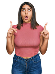 Poster - Young hispanic girl wearing casual t shirt amazed and surprised looking up and pointing with fingers and raised arms.