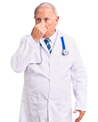 Poster - Senior handsome grey-haired man wearing doctor coat and stethoscope smelling something stinky and disgusting, intolerable smell, holding breath with fingers on nose. bad smell