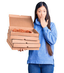 Young beautiful chinese woman holding cardboard boxes of italian pizza covering mouth with hand, shocked and afraid for mistake. surprised expression