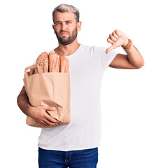 Sticker - Young handsome blond man holding paper bag with bread with angry face, negative sign showing dislike with thumbs down, rejection concept