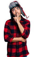 Sticker - Hispanic young man with beard wearing fluffy earmuff hat asking to be quiet with finger on lips. silence and secret concept.