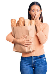 Canvas Print - Young beautiful latin girl holding paper bag with bread covering mouth with hand, shocked and afraid for mistake. surprised expression