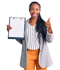 Wall Mural - Young african american woman wearing business clothes holding clipboard smiling happy and positive, thumb up doing excellent and approval sign