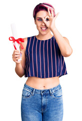 Canvas Print - Young beautiful woman with pink hair holding graduate degree diploma smiling happy doing ok sign with hand on eye looking through fingers