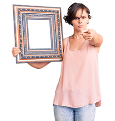 Wall Mural - Beautiful young woman with short hair holding empty frame pointing with finger to the camera and to you, confident gesture looking serious