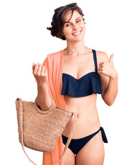 Poster - Beautiful young woman with short hair wearing bikini and hat holding summer wicker handbag smiling happy and positive, thumb up doing excellent and approval sign