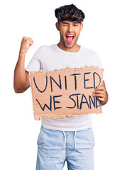 Canvas Print - Young hispanic man holding united we stand banner screaming proud, celebrating victory and success very excited with raised arms