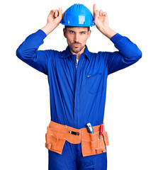 Poster - Young handsome man wearing worker uniform and hardhat doing funny gesture with finger over head as bull horns