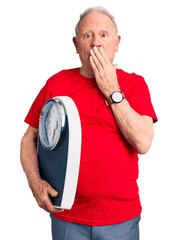 Wall Mural - Senior handsome grey-haired man holding weighing machine covering mouth with hand, shocked and afraid for mistake. surprised expression