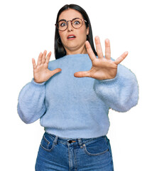 Wall Mural - Young hispanic woman wearing casual clothes and glasses afraid and terrified with fear expression stop gesture with hands, shouting in shock. panic concept.