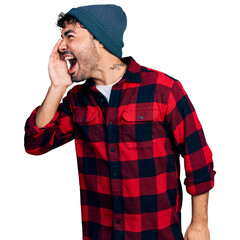 Wall Mural - Hispanic young man with beard wearing wool winter hat shouting and screaming loud to side with hand on mouth. communication concept.