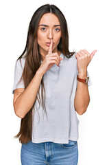 Wall Mural - Young hispanic girl wearing casual white t shirt asking to be quiet with finger on lips pointing with hand to the side. silence and secret concept.