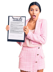 Canvas Print - Young beautiful latin girl holding clipboard with terms and conditions document scared and amazed with open mouth for surprise, disbelief face