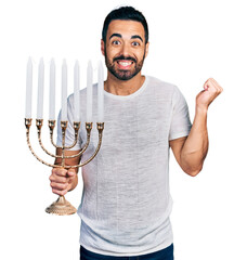 Sticker - Young hispanic man with beard holding menorah hanukkah jewish candle screaming proud, celebrating victory and success very excited with raised arm