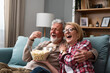 © Srdjan - Old retired age couple watching TV at home, old mature couple cheering sport games competition together with laugh smile victory on sofa couch at living room home. Senior man and woman movie night.