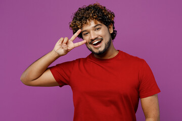 Wall Mural - Young smiling cheerful fun happy Indian man he wear red t-shirt casual clothes looking camera show cover eye with victory sign isolated on plain purple background studio portrait. Lifestyle concept.