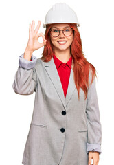 Wall Mural - Young redhead woman wearing architect hardhat smiling positive doing ok sign with hand and fingers. successful expression.