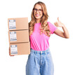 © Krakenimages.com - Young caucasian woman with blond hair holding delivery package smiling happy and positive, thumb up doing excellent and approval sign