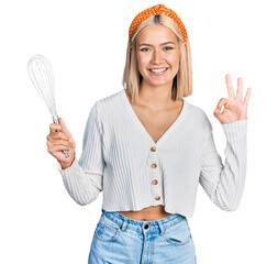 Wall Mural - Beautiful young blonde woman holding baker whisk doing ok sign with fingers, smiling friendly gesturing excellent symbol