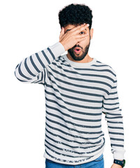 Sticker - Young arab man with beard wearing casual striped sweater peeking in shock covering face and eyes with hand, looking through fingers with embarrassed expression.