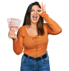 Wall Mural - Beautiful hispanic woman holding 10 colombian pesos banknotes smiling happy doing ok sign with hand on eye looking through fingers