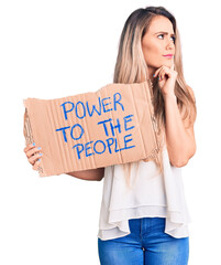 Wall Mural - Young beautiful blonde woman holding power to the people cardboard banner serious face thinking about question with hand on chin, thoughtful about confusing idea