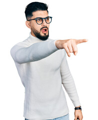 Wall Mural - Young arab man with beard wearing elegant turtleneck sweater and glasses pointing with finger surprised ahead, open mouth amazed expression, something on the front