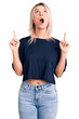 Young beautiful blonde woman wearing casual t-shirt amazed and surprised looking up and pointing with fingers and raised arms.