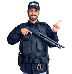 Canvas Print - Young hispanic man wearing police uniform holding shotgun smiling happy pointing with hand and finger to the side