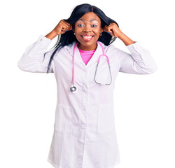 Poster - Young african american woman wearing doctor stethoscope smiling pulling ears with fingers, funny gesture. audition problem