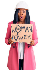 Poster - Young african american woman wearing architect helmet holding woman power banner scared and amazed with open mouth for surprise, disbelief face